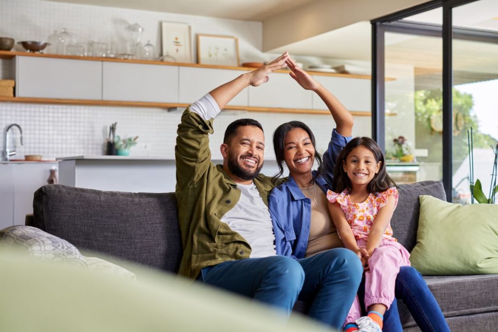 Happy family sitting on a couch at home, symbolizing the peace of mind and support provided by transaction coordination services during the home buying or selling process. The parents form a roof shape with their arms over their heads, representing security and a smooth real estate experience, while their smiling daughter sits between them